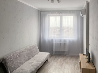 A simple room interior with a gray silver sofa with a sideboard and a TV opposite, with gray textured walls and white tulle on the window