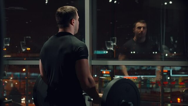 Muscular man in black sportswear standing against window and exercising with barbell, his reflection in glass, lights of night city lying beneath. Bodybuilder from behind working out in gym