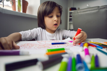 boy child sits at home at a table and draws with colored markers on a white sheet