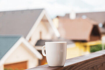 The steam rises from a cup of hot coffee or tea on the balcony with mountains and roofs of houses on the background. 