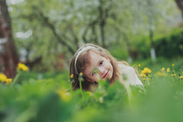 Naklejka na ściany i meble A beautiful girl in white hiding in grass in spring garden. Portrait of child among green grass and yellow dandelions. Young lady sitting in sunny blooming park and smilling.