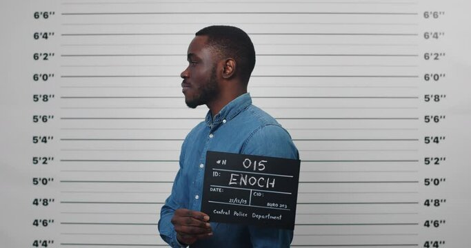 Mugshot of afro american guy in 30s turning to sides while holding sign and posing for photo . Crop view of young man looking to camera in police department. Concept of crime.