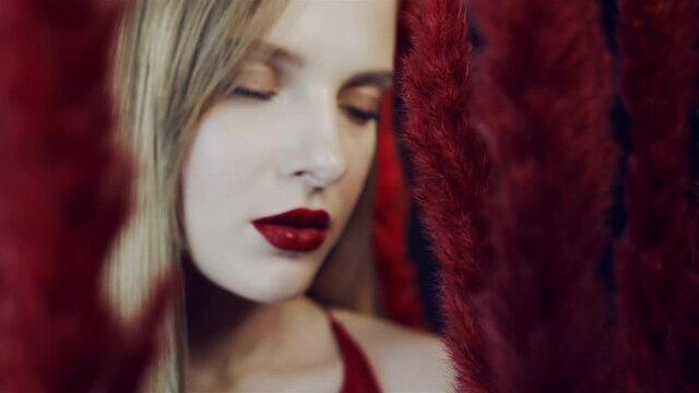 Face of a young woman with red lipstick among red fluffy feathers. The girl looks at the camera. The girl among the racist exotic plants. Blonde with red lips. Camera zooms in on the face.