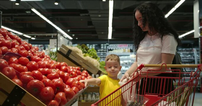 Cute woman with her little happy son chooses fresh vegetables together at the grocery store. A mother with a baby boy takes ripe tomatoes from the shelf and puts them in a shopping basket.