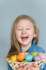Fototapeta na wymiar A beautiful blond girl, four years old, in a denim dress, hold in her hand basket with colored Easter eggs. A girl stands on a gray background, smile, has fun, rejoices.