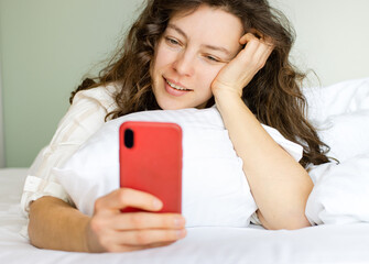 Beautiful young woman using smartphone, lying in white cozy bed at home. Checking social media,...