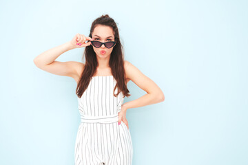 Young beautiful smiling female in trendy summer hipster overalls clothes. Sexy carefree woman posing near light blue wall in studio. Positive model having fun indoors. Makes duck face in sunglasses