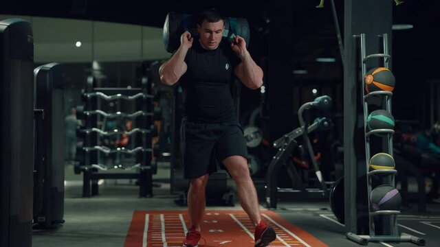 Bodybuilder in black sportswear doing alternate lunges exercise with sand bag on shoulder, unrecognizable people on background. Tracking shot athlete training in gym. Concept of sport