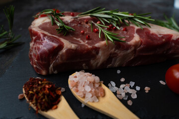 Close-up piece of beef meat ribeye with spoons of Himalayan salt and grilling spices, tomato and rosemary. Side view. BBQ ingredients.