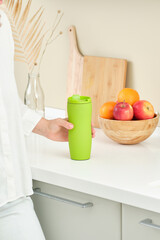 Woman holdin a green thermo mug on the background of a wooden cup with oranges and apples. Healthy food concept. Reusable eco-friendly kitchen products. Zero waste and sustainable plastic free