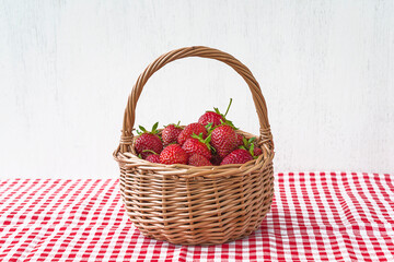 Fototapeta na wymiar Wicker basket with ripe strawberries on checkered tablecloth and white paint wooden background