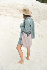 Young girl in natural linen clothes and a straw hat, barefoot on a white sand beach, holding a linen bag in her hands. Side view.