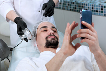 Man makes facial skin care in a beauty salon. Men's cosmetology. The man holds the phone in his...