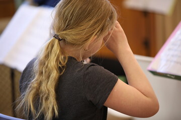 Portrait of a little girl with long hair gathered in a ponytail sits with a raised hand at her head and touches her forehead at a music lesson in class with notes.Tired child.Back view.Close-up