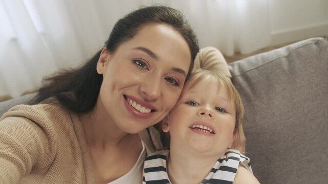 Young mother and small preschooler daughter look at camera making selfie and smiling at home together. Happy funny mom and little girl child have fun laugh take photo posing for self-portrait picture