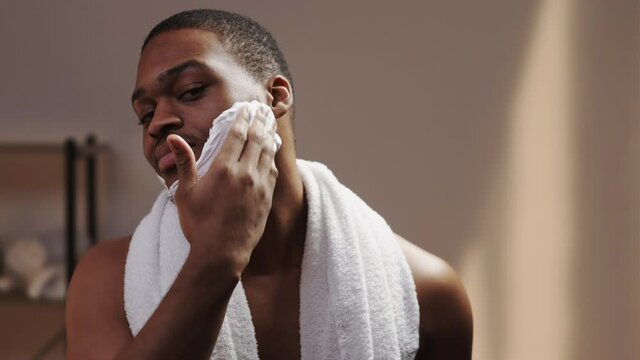 Male skincare. Cosmetic product. Grooming routine. Confident shirtless athletic African man with towel applying shaving foam on face in bathroom on brown wall background.