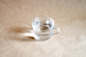 Obraz na płótnie Canvas a glass cup of clear water on beige craft paper background and reflection from the light