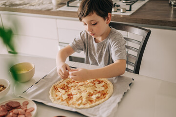 Little kid boy making pizza sitting at the table on the kitchen. Children helping in cooking lifestyle image