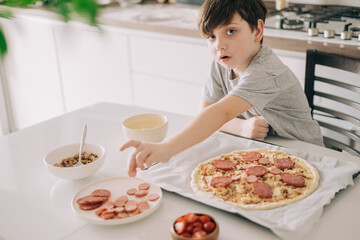 Little kid boy making pizza sitting at the table on the kitchen. Children helping in cooking lifestyle image