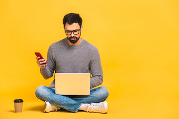 Young bearded man sitting on the floor with laptop and talking at phone. Isolated over yellow background. - 425878926