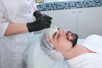 Beautician applies a nourishing mask to the man's face. A man doing facial procedures in a cosmetology clinic. Male cosmetology. The concept of preservation of youth and skin care of the face.