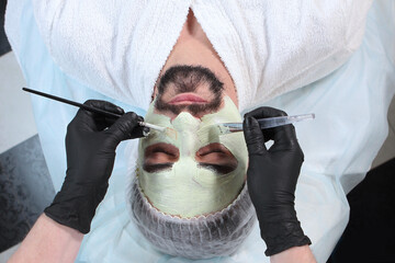 Beautician applies a nourishing mask to the man's face. A man doing facial procedures in a cosmetology clinic. Men's cosmetology. 