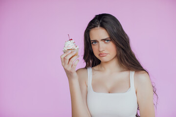 Sad and heartbroken lonely young girl broke-up with boyfriend crying and sobbing, holding cake, eating to ease pain, standing sulky and unhappy over pink background, express sadness and distress