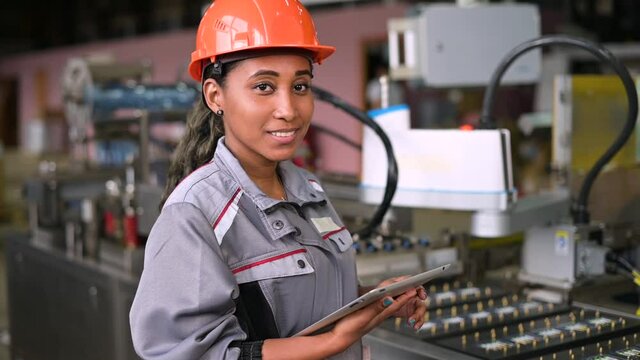 Slow motion Portrait of a beautiful smiling black factory worker. Young woman in an orange industrial hard hat with a tablet in her hands controls production at a factory