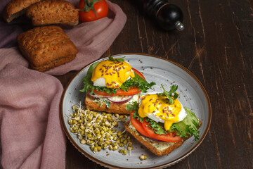 Eggs benedict with mustard sauce on a grain bun, with fresh vegetables and cream cheese. Close-up, on a dark wooden surface.