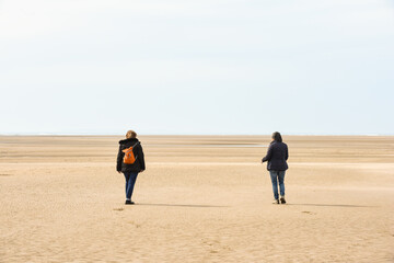 Two friends meet and go for a walk on the beach at low tide whilst keeping a safe distance apart in accordance with coronavirus restriction rules in England