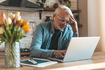 Financial reports and statistics. Concentrated senior man working on laptop at home in the kitchen....