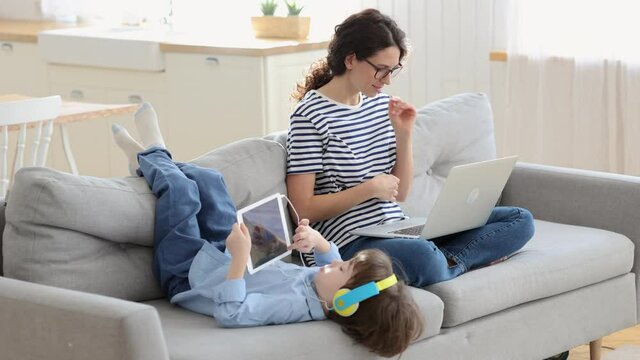 Family, distance job concept. Happy mother work from home on laptop during lockdown, sitting on couch, distracted by son and tickles belly, kid playing on digital tablet wear headphones. 