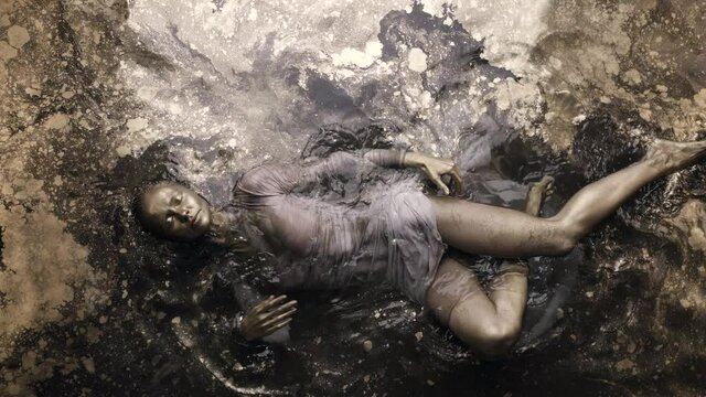 A woman in a dress with dyed golden skin and hair lies in the water, slowly moving and posing.