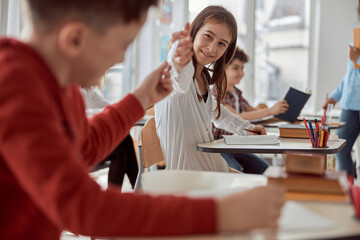 Schoolboy gives pencil to his classmate while sitting at desk while teacher speaking in school classroom