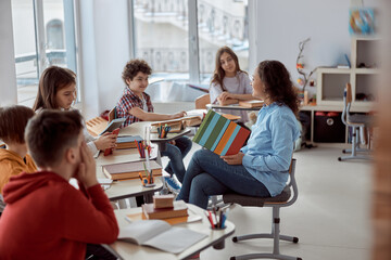 Young tutor is teaching to read her student. Elementary school kids sitting on desks and reading...