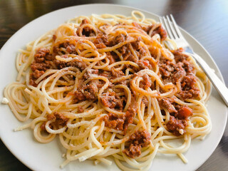Spaghetti Bolognese with minced beef, onion, chopped tomato, garlic, olive oil, stock cube, tomato puree and Italian herb. Traditional Italian food on a white plate with a fork on the right. 