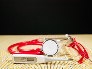 White electric thermometer in focus, red stethoscope and red glasses out of focus on a a bamboo...