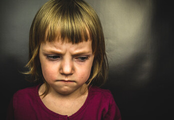 Little blonde girl on a black background with eyes down. Emotion of pain.The concept of abuse of children and the theme of raising children