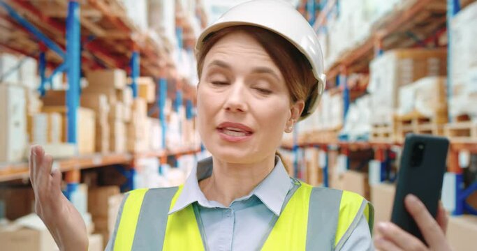 Beautiful female warehouse worker in white helmet talks on video call using smartphone while standing on background of rows with parcels.