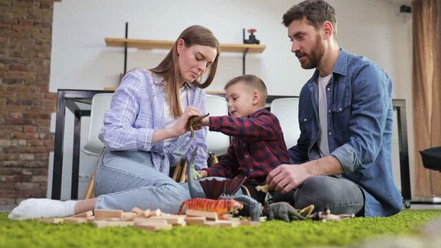 Happy young mom and dad playing game with little son sitting on green floor holding dinosaurs having fun with toys and wooden blocks at home