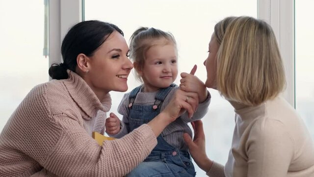 Two mothers and a young daughter spend time at home. They're playing on the window sill. Warm home clothes. Lgbt family, lesbians with a child.