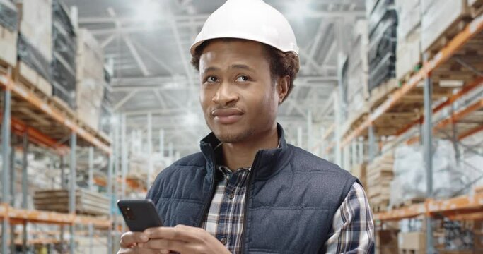 Happy handsome young African American man worker builder in helmet standing at factory working and speaking on cellphone. Male professional constructor talking on smartphone, close up concept