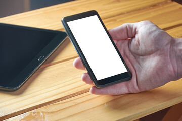 A young adult man's hand holding black mobile phone with white empty screen on rustic wooden table. Technology, communications, working from home.