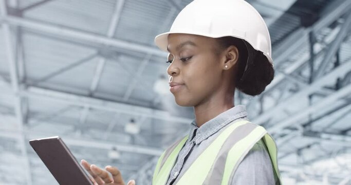 African American young female builder in helmet standing at factory working using tablet typing online. Side view of pretty woman engineer browsing on device making checklist at factory, close up