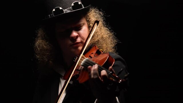 Portrait of man in an original hat in Irish style plays violin on black background. Violinist masterfully manipulates violin bow, playing strings in dark studio, illuminated by bright lights. Close up