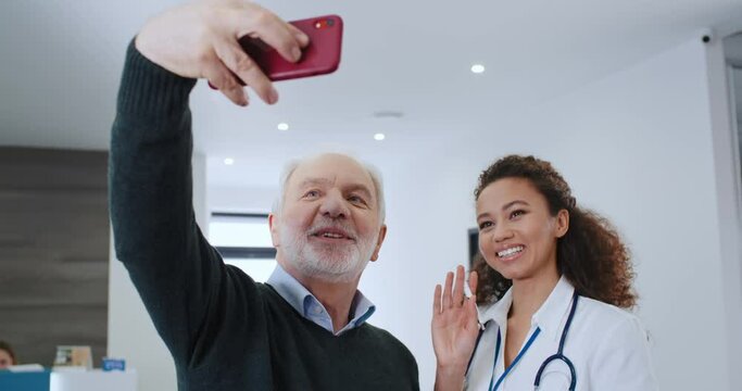 Young african american doctor in medical coat and gray-haired senior patient with beard making selfie photo with smartphone in hospital. Mixed race nurse and happy elderly caucasian man