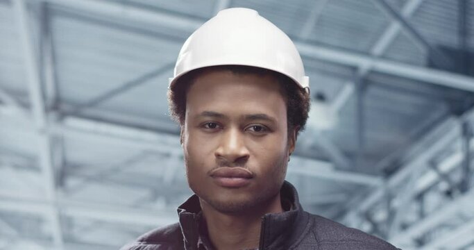 Close up shot of cheerful African American young man engineer specialist looking at camera standing at factory in white helmet, looking at camera and smiling, Handsome builder with smile on face