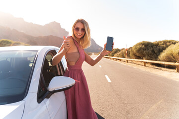 Naklejka premium Happy blonde woman showing smartphone and showing thumb up, standing near the rental car.