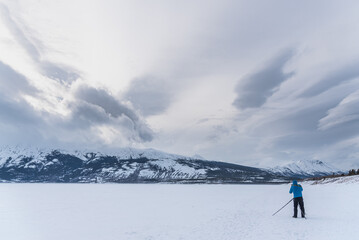 Man in a blue jacket standing on a frozen lake photographing, taking photos of the stunning, snowy, winter landscape with clouds shining above. 