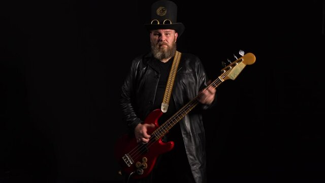 Portrait of man in leather coat and an original hat playing bass guitar. Rock musician dynamically plays strings of an instrument on black studio background. Live rock concert, heavy metal. Close up.